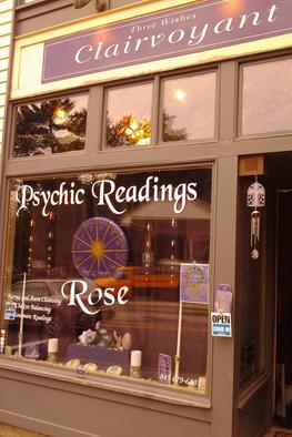 A photo of the storefront of Rose's shop on the main street in Woodstock. Specific address of Psychic Readings by Rose is 40 Mill Hill Rd. Woodstock, New York, 12498. This photo was taken in the summer of 2012. The store's banner atop states "Three Wishes Clairvoyant" on a dark purple sign. The glass windows of the store has "Psychic Readings Rose" in white font. There's a hand painted purple and gold astrology wheel hanging in the middle of window.
